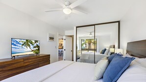 Une chambre d’hôtel moderne avec un grand lit, un téléviseur posé sur une commode en bois, un ventilateur de plafond et une porte-fenêtre coulissante donnant sur un balcon avec vue sur les palmiers.