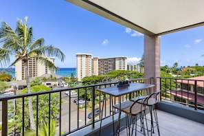 Varanda com mesa e cadeiras, vista para uma rua com carros e edifícios, incluindo um hotel alto e o oceano.