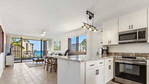 Une cuisine moderne avec des meubles blancs, un grand îlot central, des appareils électroménagers en acier inoxydable et une vue sur les palmiers à travers de grandes fenêtres.