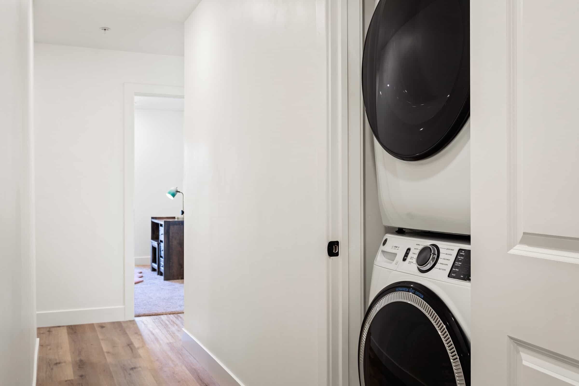 A laundry room with a front-loading washer and dryer, wooden flooring, and a hallway leading to another room.