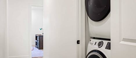 A laundry room with a front-loading washer and dryer, wooden flooring, and a hallway leading to another room.