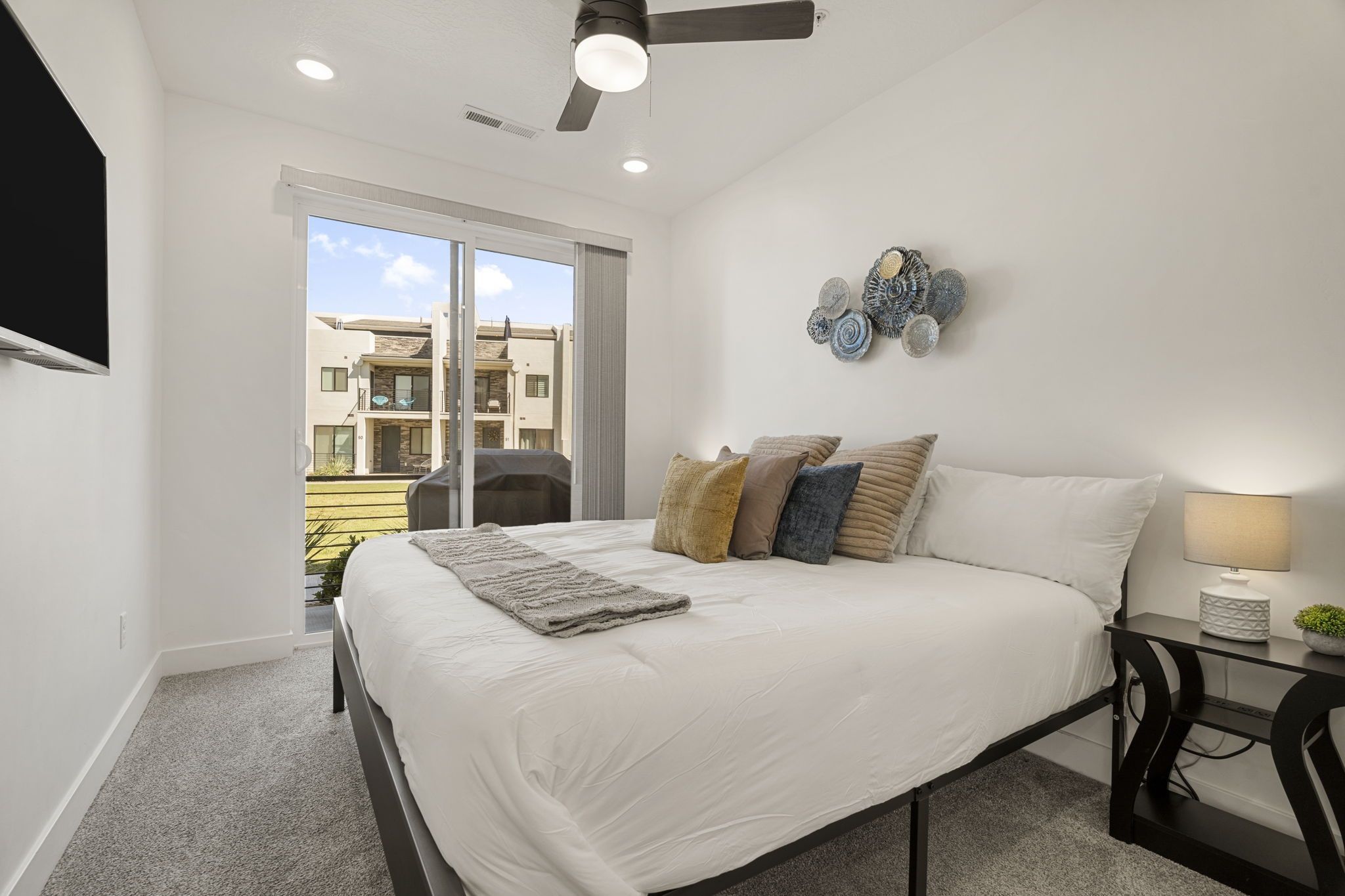 A bedroom with a bed, a ceiling fan, a wall-mounted TV, and a view of the outside.