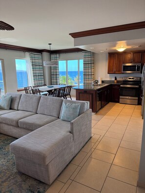 A modern living room with a large sectional sofa, a dining area with a view of the ocean, and a kitchen with wooden cabinets and stainless steel appliances. - World Class Resort Marriott Maui Ocean Club Ground Floor Ocean Front 3 bd 3bth (Lahaina)