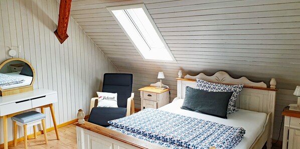 A bedroom with a sloped ceiling, a bed with patterned bedding, a desk with a chair, and a small table with a lamp. - Lovingly guest apartments Alpirsbach - Apartment 3 (Alpirsbach)