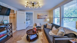 A living room with a sofa, a coffee table, a TV, and a kitchen area visible through an open door. - Oak - Walk to the Stadium!  Backyard with firepit & grill (Tuscaloosa)