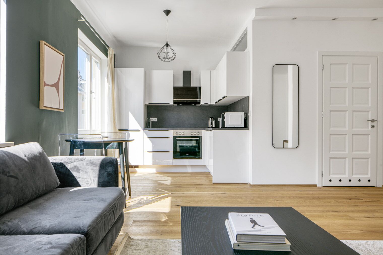 A modern living room with a grey sofa, a coffee table, and a book titled 'KIMTOLK'. A kitchen with white cabinets, a black countertop, and a microwave is visible in the background.