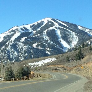 A winding mountain road with a guardrail, surrounded by snow-covered slopes and sparse trees.