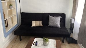 Un sofá negro con dos cojines, una mesa blanca con una planta y un control remoto, un cuadro de un paisaje urbano y una lámpara sobre una mesita auxiliar.