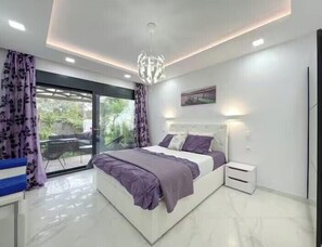 Um quarto moderno com uma cama grande, uma mesa de cabeceira, uma porta de vidro que dá para uma varanda e um lustre no teto.