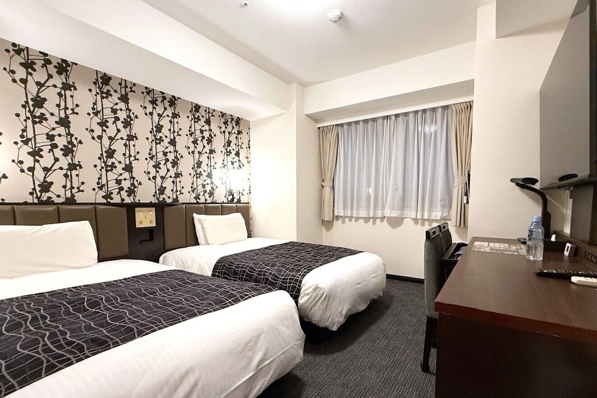 A hotel room with two beds, a desk, a mirror, a window with curtains, and a wall with a floral pattern.