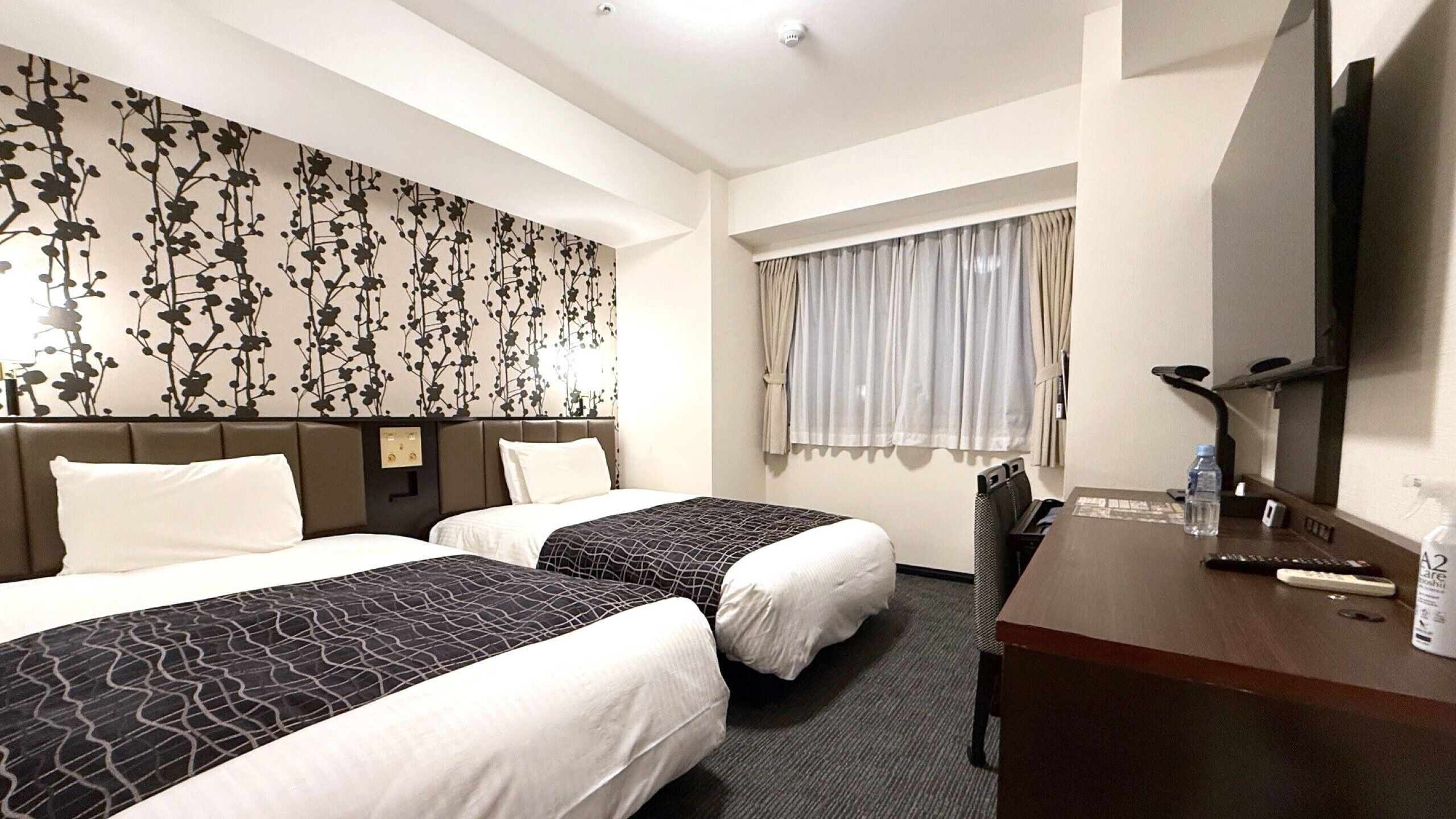 A hotel room with two beds, a desk, a television, and a window with curtains.