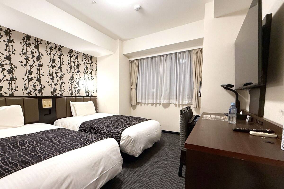 A hotel room with two beds, a desk, a television, and a window with curtains.
