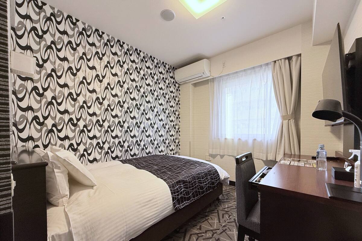 A hotel room with a patterned wall, a bed with a patterned bedspread, a desk with a lamp, and a window with curtains.