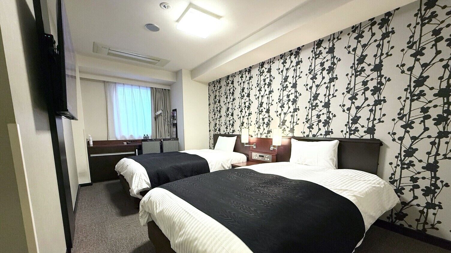 A hotel room with two beds, a TV, a desk, a chair, a window with curtains, and a floral wallpaper.