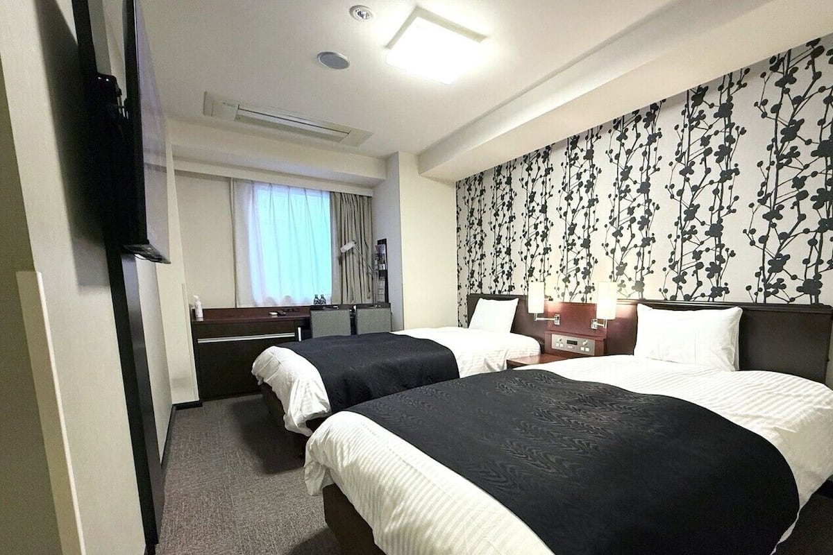 A hotel room with two beds, a TV, a desk, a chair, a window with curtains, and a floral wallpaper.