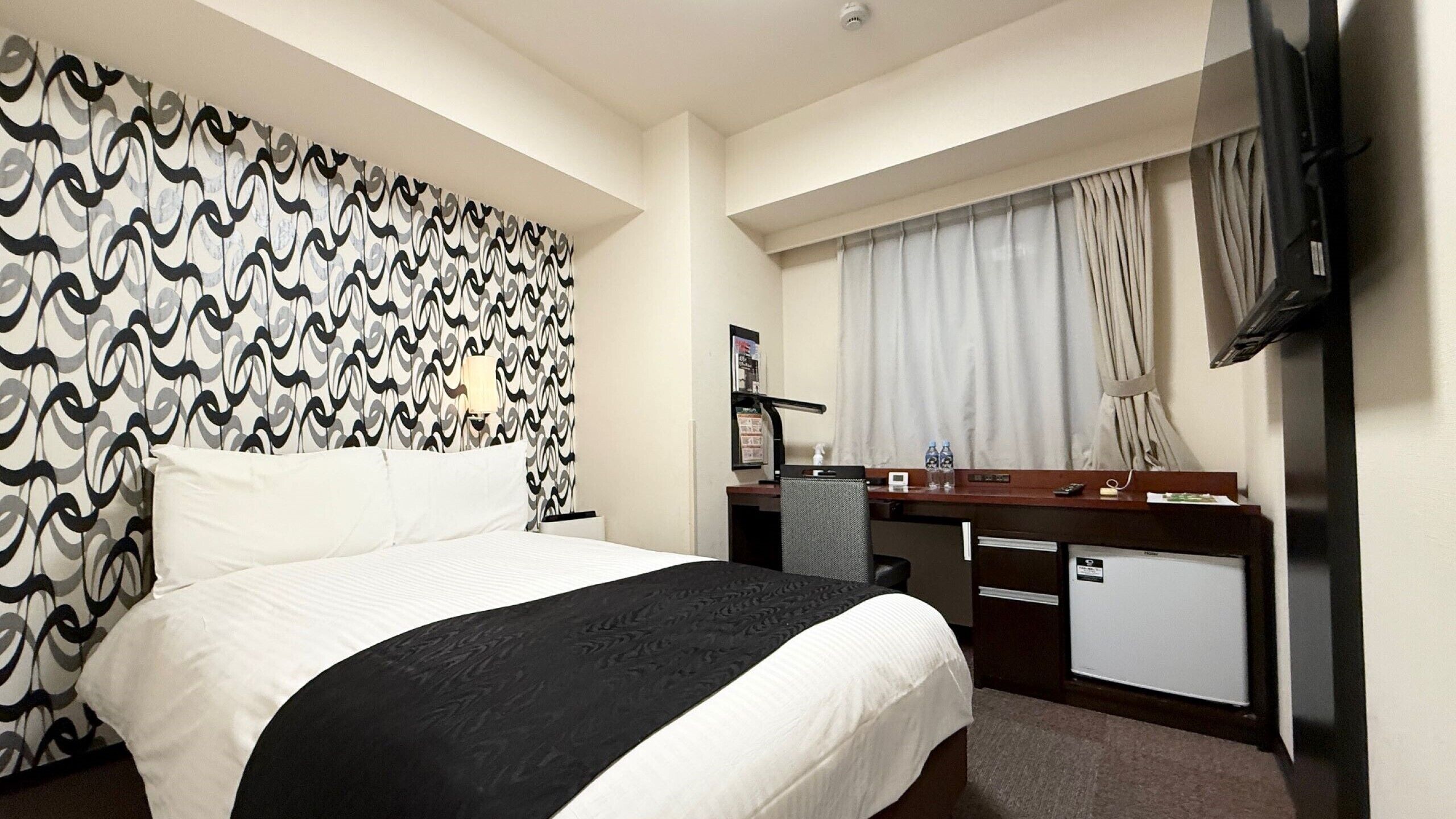 A hotel room with a bed, a desk, a chair, a TV, and a window with curtains.