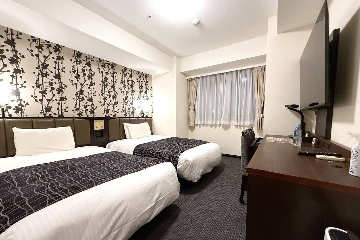 A hotel room with two beds, a desk, a TV, and a window with curtains.