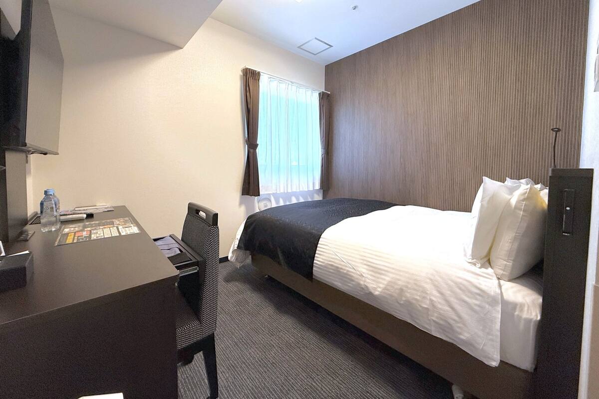 A hotel room with a bed, a desk with a chair, a TV, and a window with curtains.