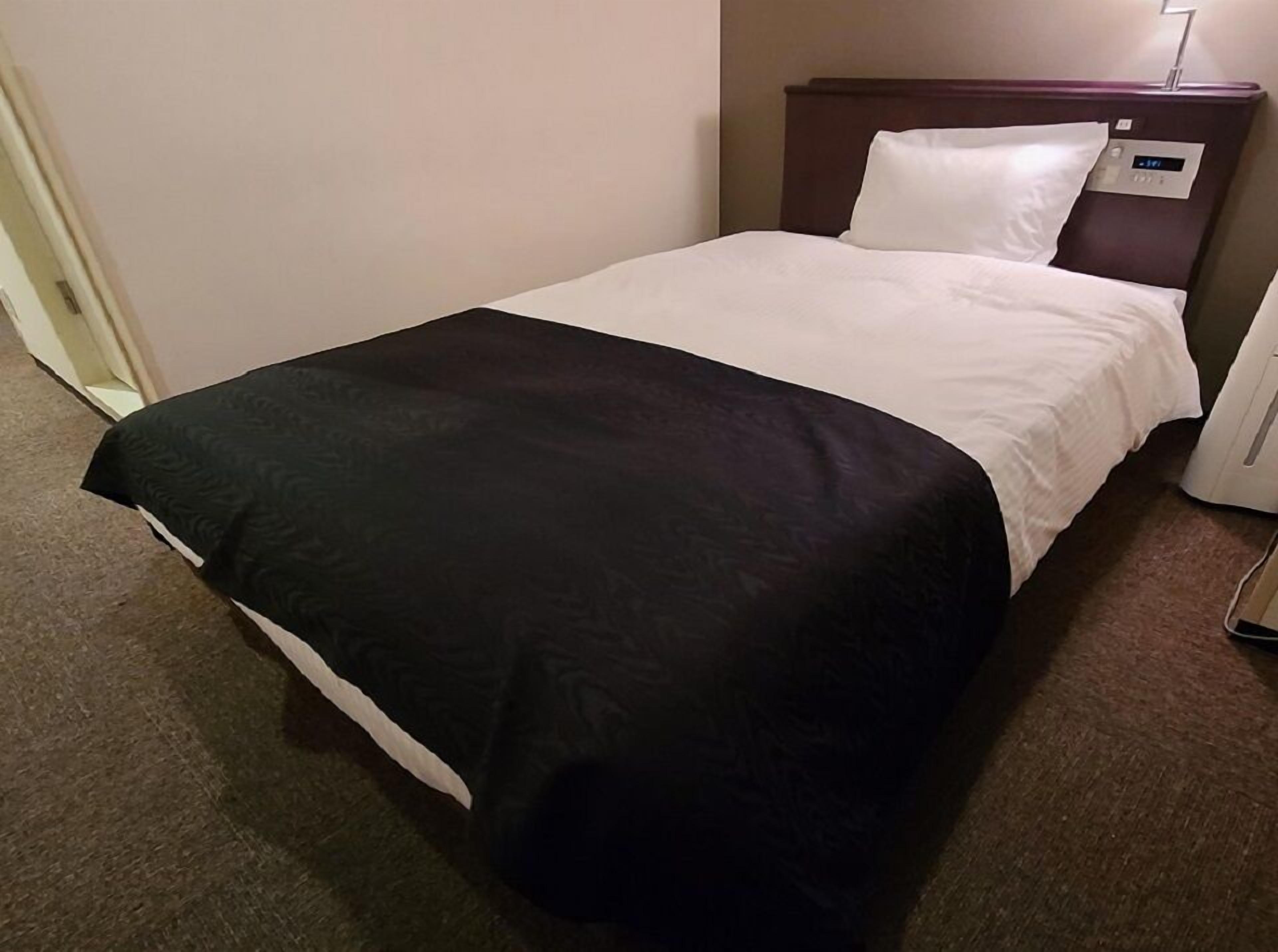 A hotel room with a bed, a bedside table, and a wall-mounted control panel.