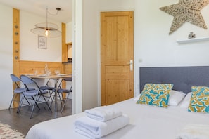 Un dormitorio con cama, almohadas, una mesita de noche, una puerta de madera y un comedor con sillas y una mesa.