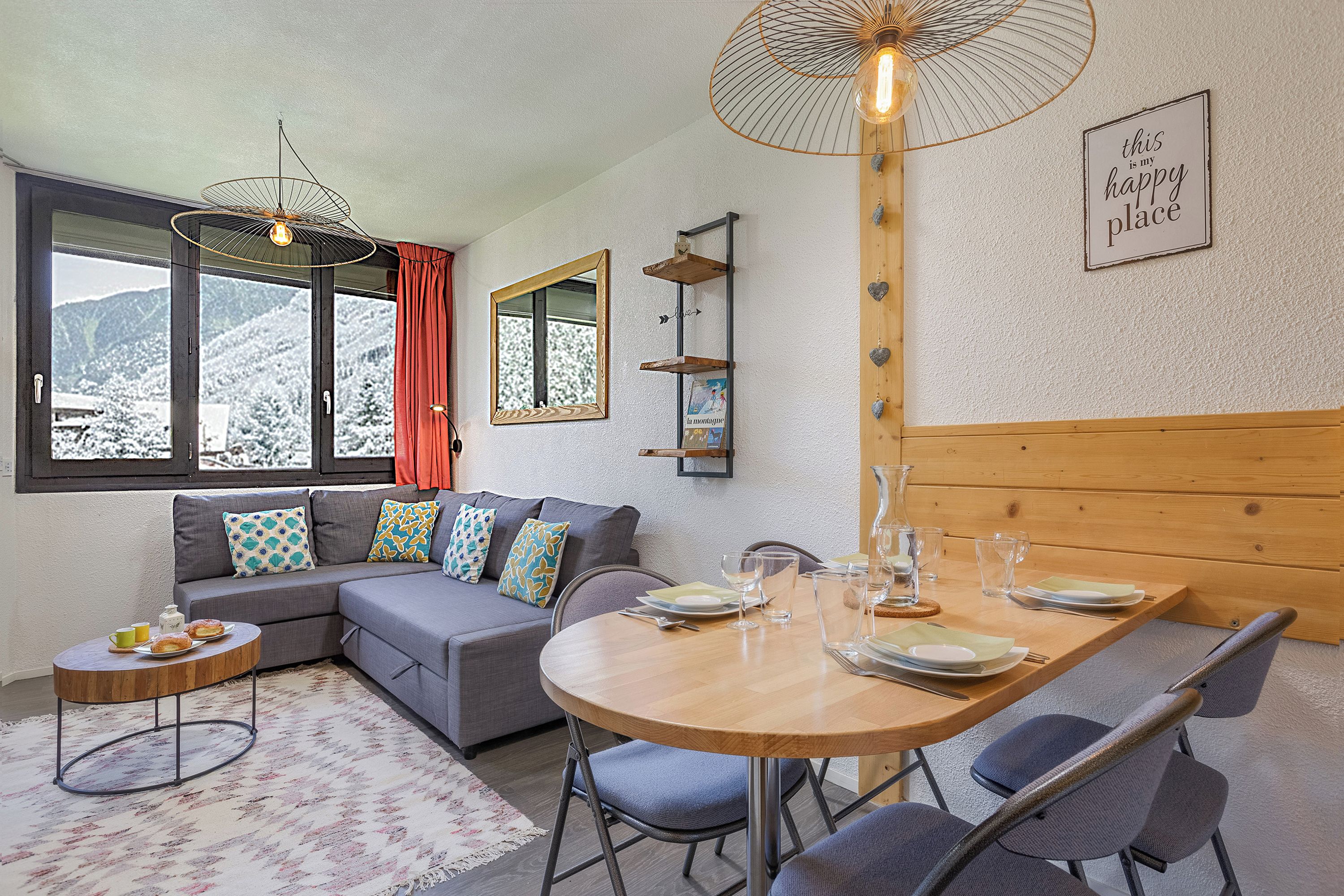 A cozy living room with a sofa, a dining table set for two, and a large window with a view of snow-covered mountains.
