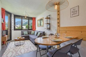 A modern living room with a sofa, dining table, and chairs, large windows with curtains, and a view of mountains. - Chamonix Sud - Chamois Blanc 512, Chamonix, France (Chamonix)