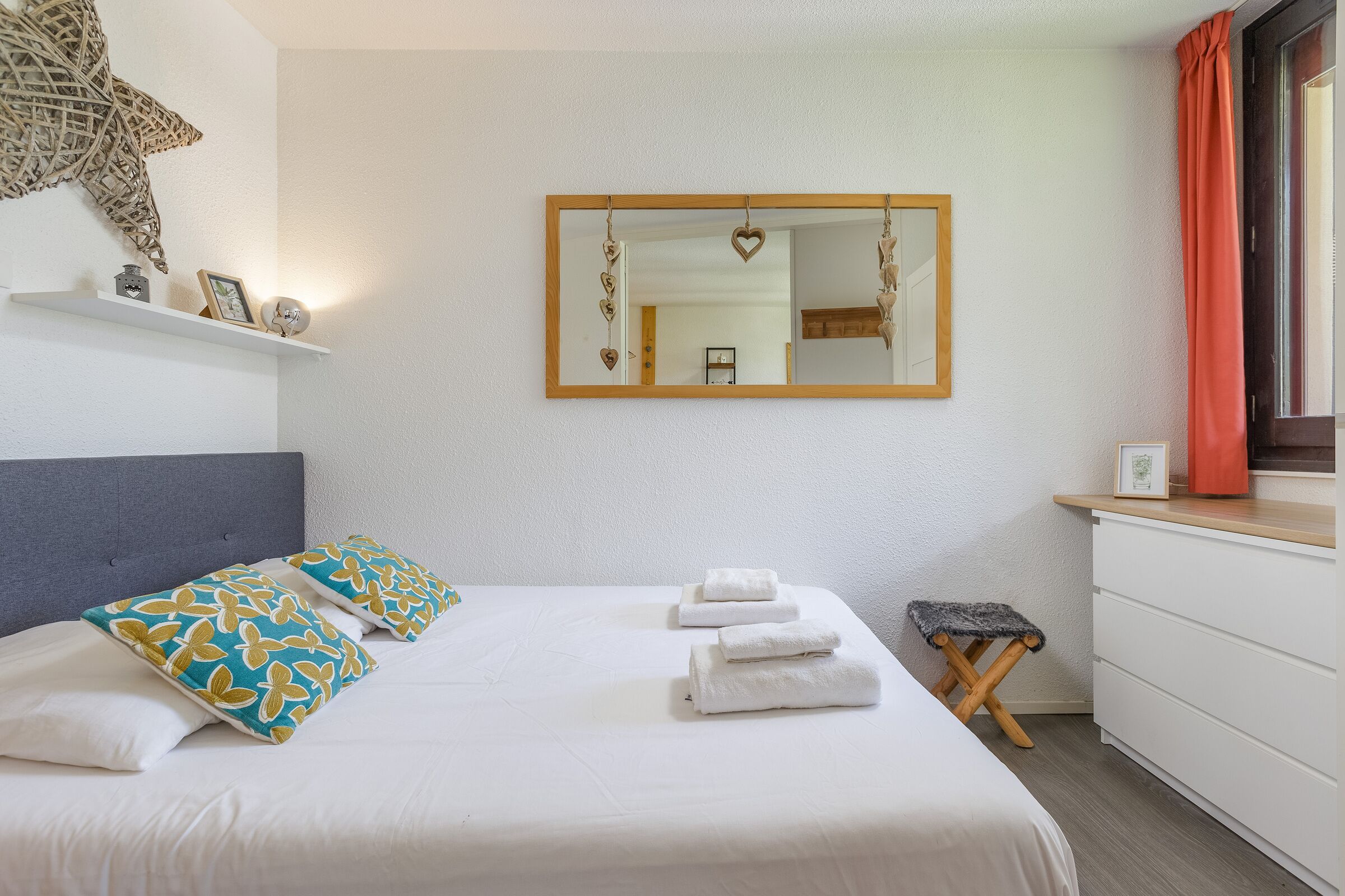 A neatly made bed with white linens and patterned pillows, a bedside table with a framed picture, a small stool, and a mirror with heart-shaped decorations.
