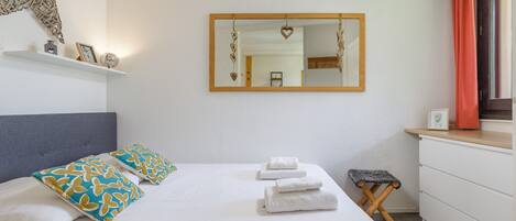 A neatly made bed with white linens and patterned pillows, a bedside table with a framed picture, a small stool, and a mirror with heart-shaped decorations.