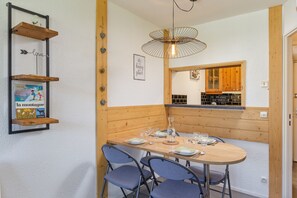 Un comedor con una mesa de madera dispuesta para cuatro personas, rodeada de sillas azules. Una estantería de madera con adornos y un cuadro están colocados en la pared.