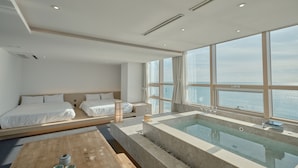 Une chambre d’hôtel moderne avec un grand lit, une table en bois sur laquelle sont posées des théières, et un jacuzzi en verre donnant sur la mer.