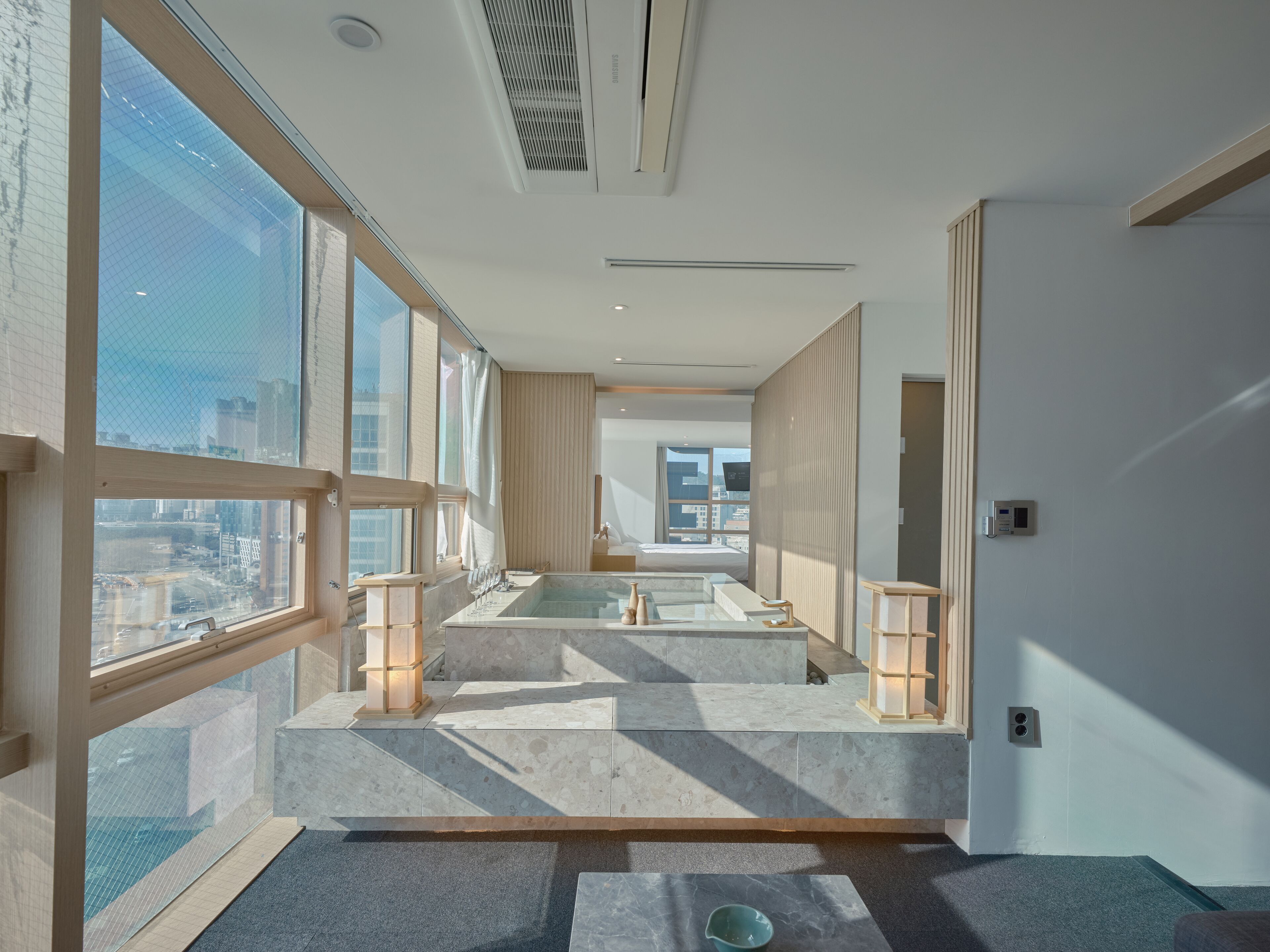 A modern hotel room with large windows, a bathtub, and a view of the city.
