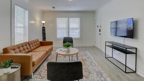 Un salon moderne avec un canapé, une table basse ronde ornée d’une plante, un fauteuil noir, un téléviseur posé sur un meuble et un tapis à motifs.