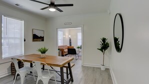 Un espace repas moderne avec une table en bois et des chaises blanches, un ventilateur de plafond et une vue sur une autre pièce où se trouvent un canapé et une lampe.