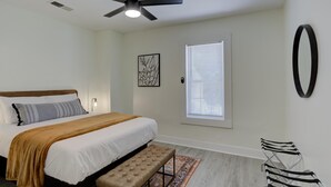 Une chambre à coucher comprenant un lit, un ventilateur de plafond, une fenêtre avec des stores, une œuvre d’art accrochée au mur et un banc.