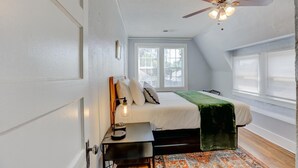 A bedroom with a bed, a desk, a chair, a ceiling fan, and a window.