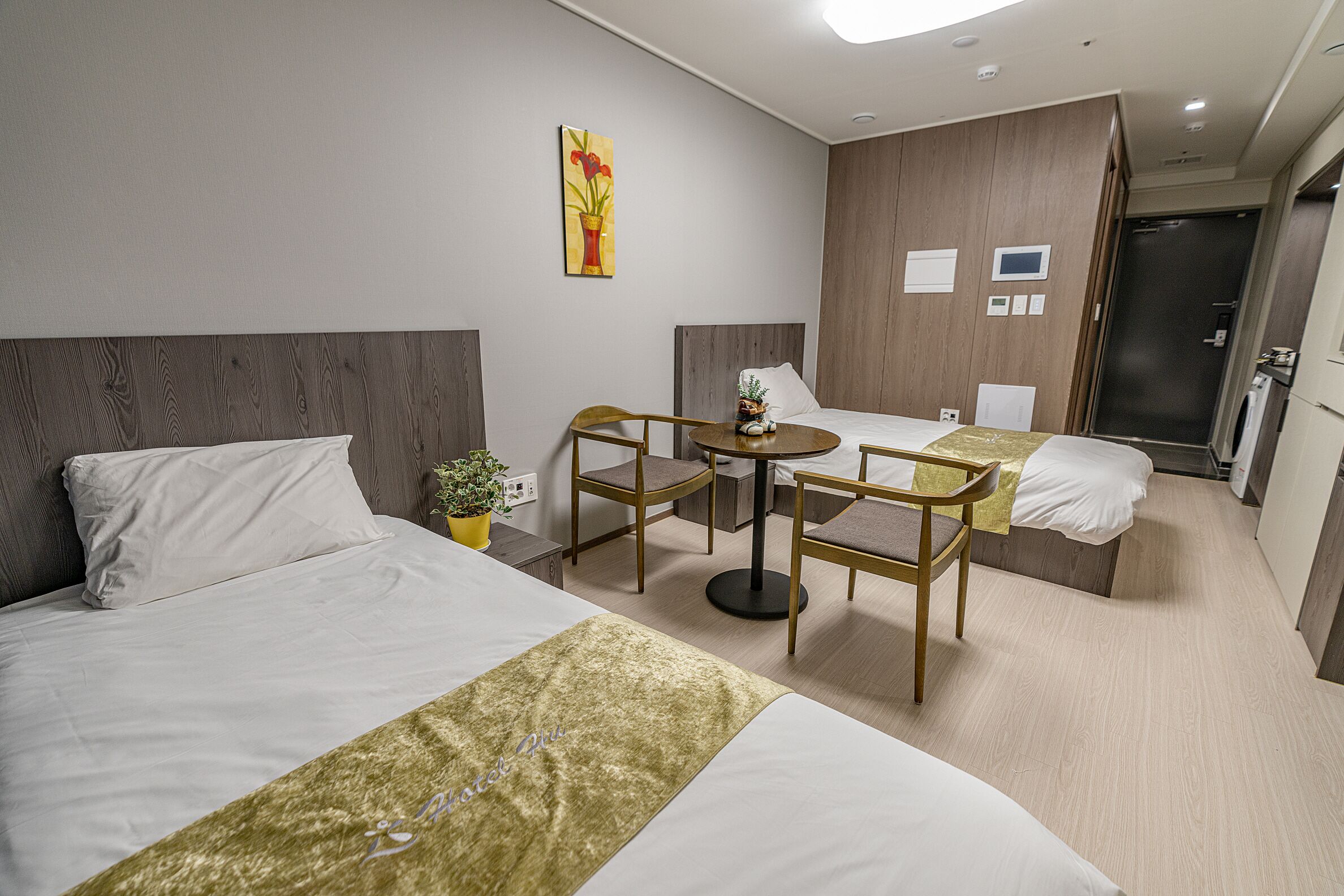 A hotel room with two beds, a small table, and a chair. There is a painting on the wall, a potted plant, and a door leading to the bathroom.