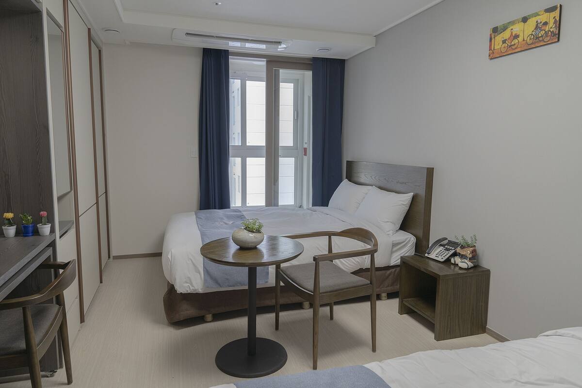 A hotel room with a bed, a small table with a plant, a chair, a bedside table with a phone, and a window with curtains.