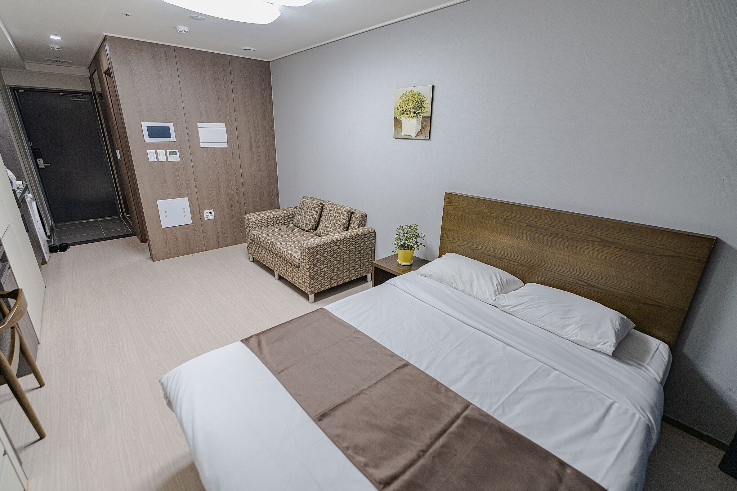 A modern hotel room with a bed, a chair, a bedside table, a wall-mounted TV, and a door.