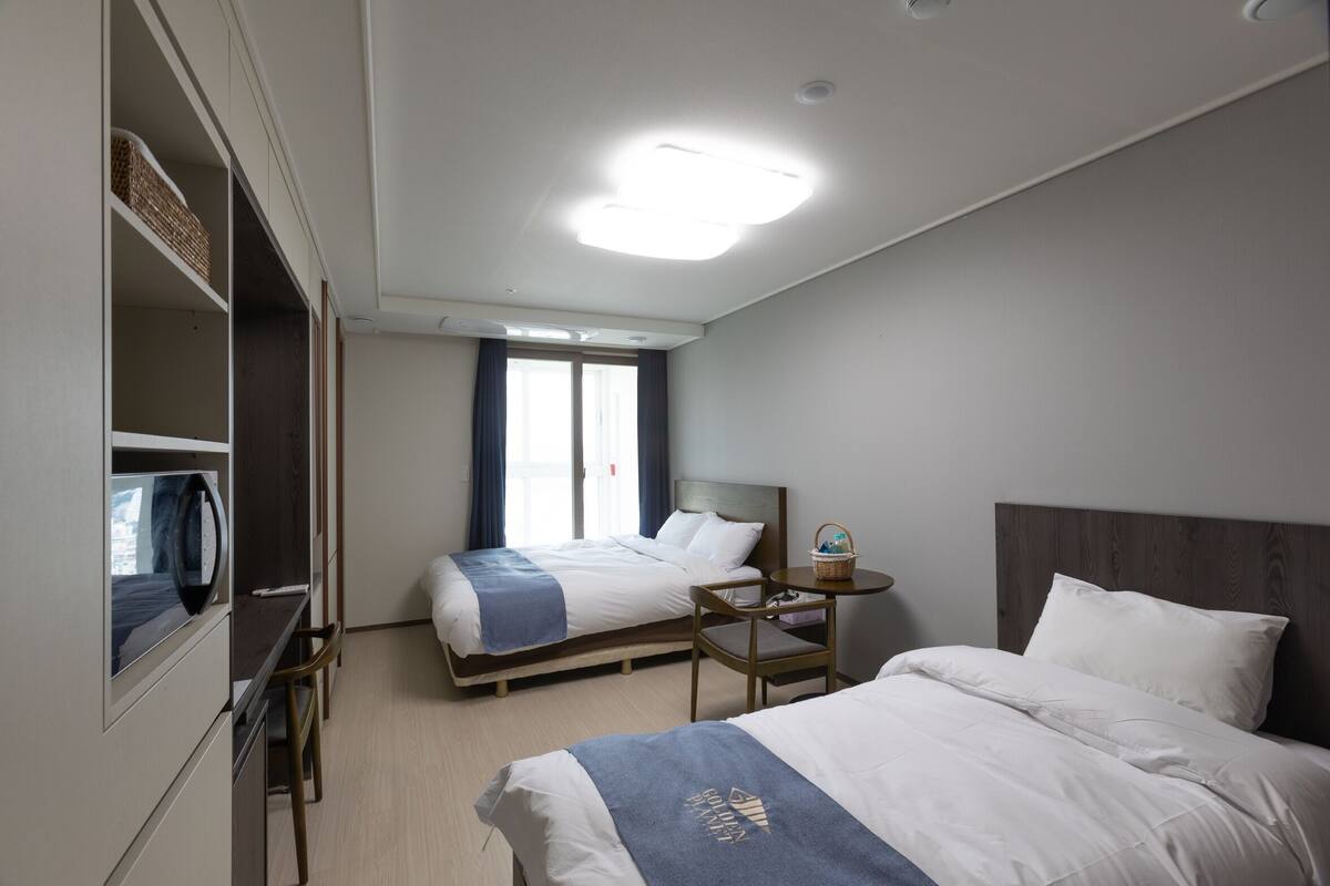 A hotel room with two beds, a desk, a chair, a small table with a basket, and a window with curtains.