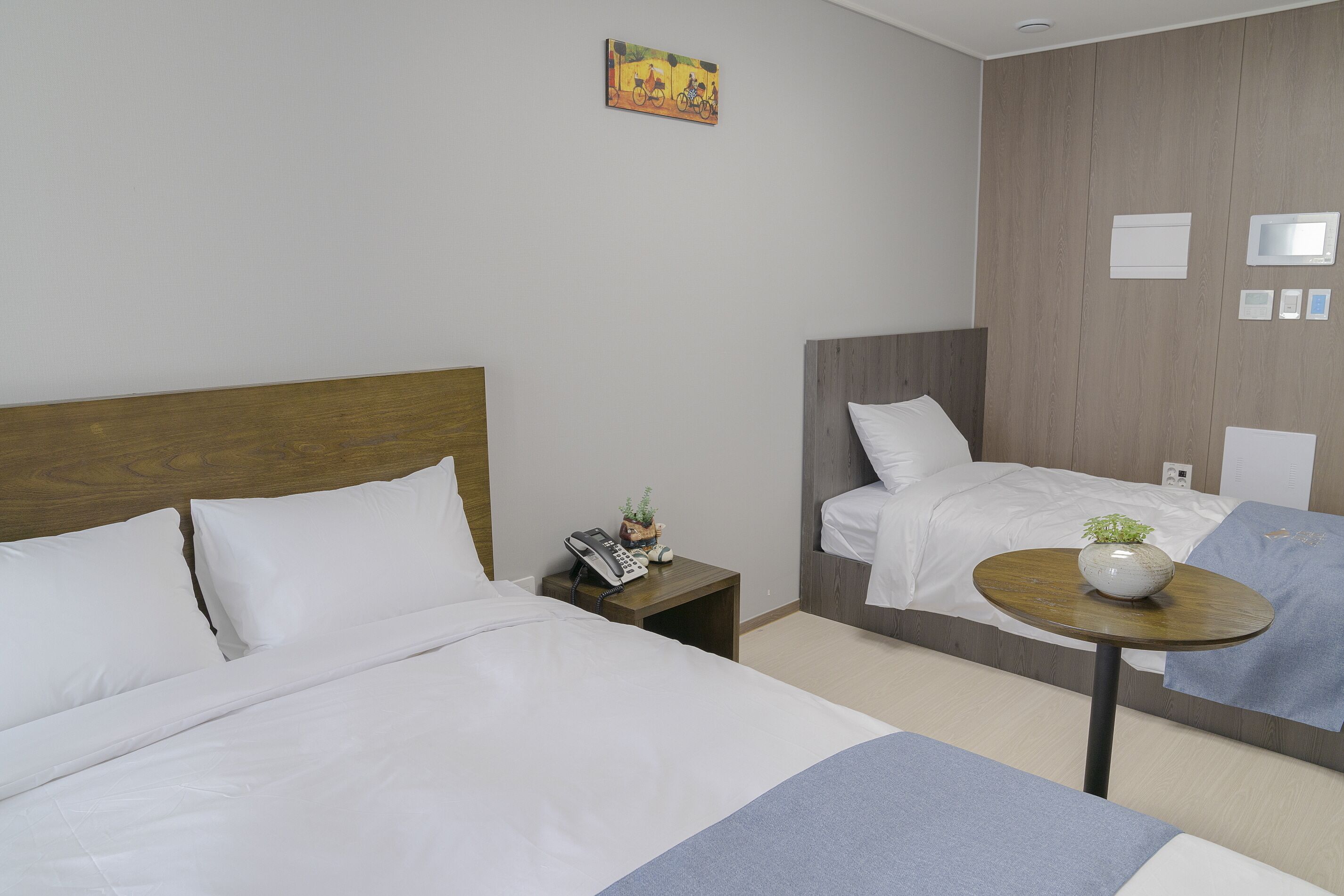 A hotel room with a large bed, a bedside table with a phone and a small plant, a round table with a vase, and a wall-mounted control panel.