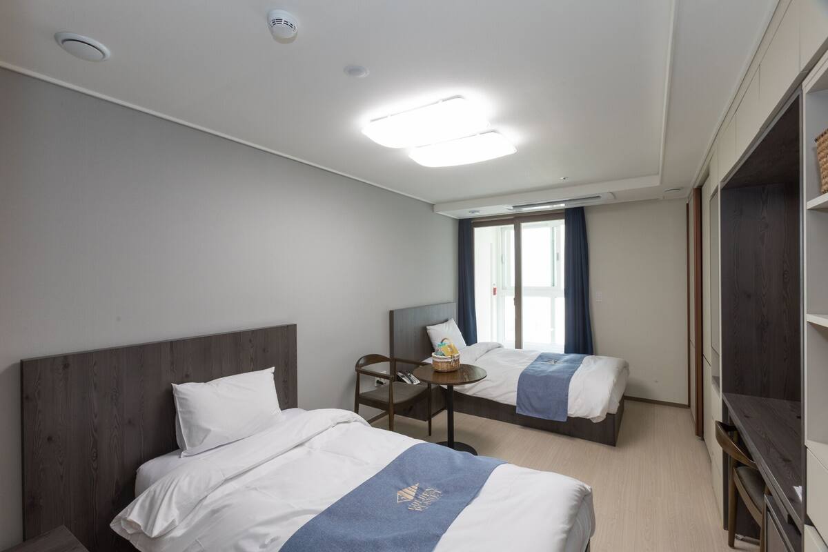 A hotel room with two beds, a small table, a chair, a wardrobe, and a window with curtains.