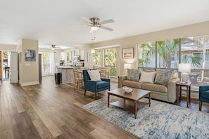 A spacious living area with a kitchen, a dining area, and large windows with a view of palm trees.