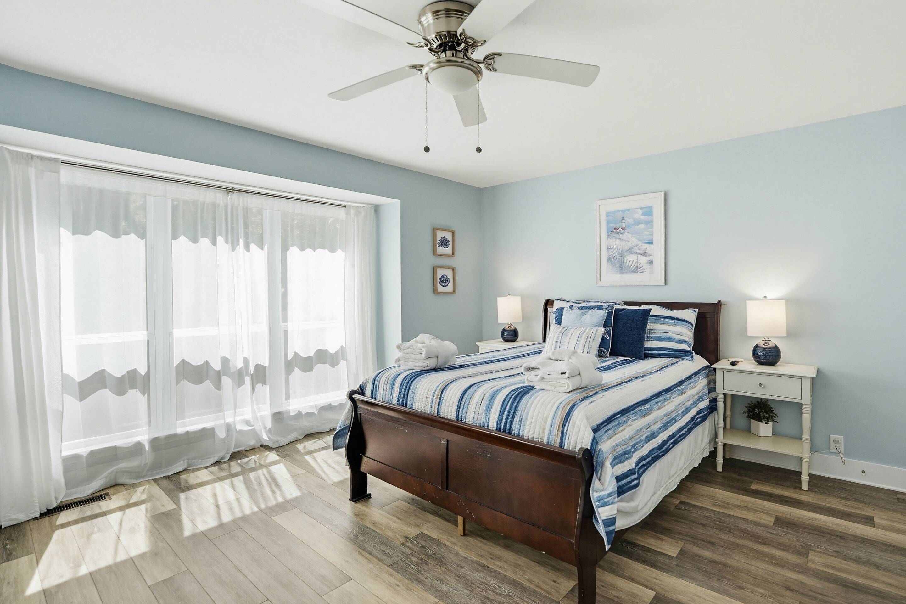 A bedroom with a bed, a ceiling fan, a nightstand, a lamp, and a window with sheer curtains.