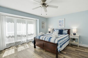 Een slaapkamer met een bed, een plafondventilator, een nachtkastje, een lamp en een raam met dunne gordijnen.