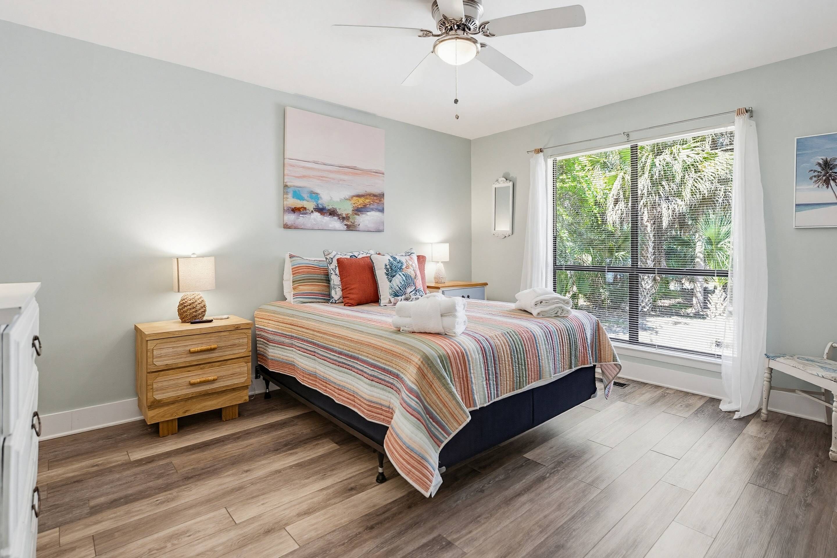 A bedroom with a bed, a nightstand, a ceiling fan, a window with blinds, and a painting on the wall.