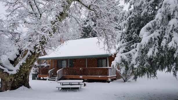 Een chalet met een houten terras en een overdekte veranda, omgeven door met sneeuw bedekte bomen.