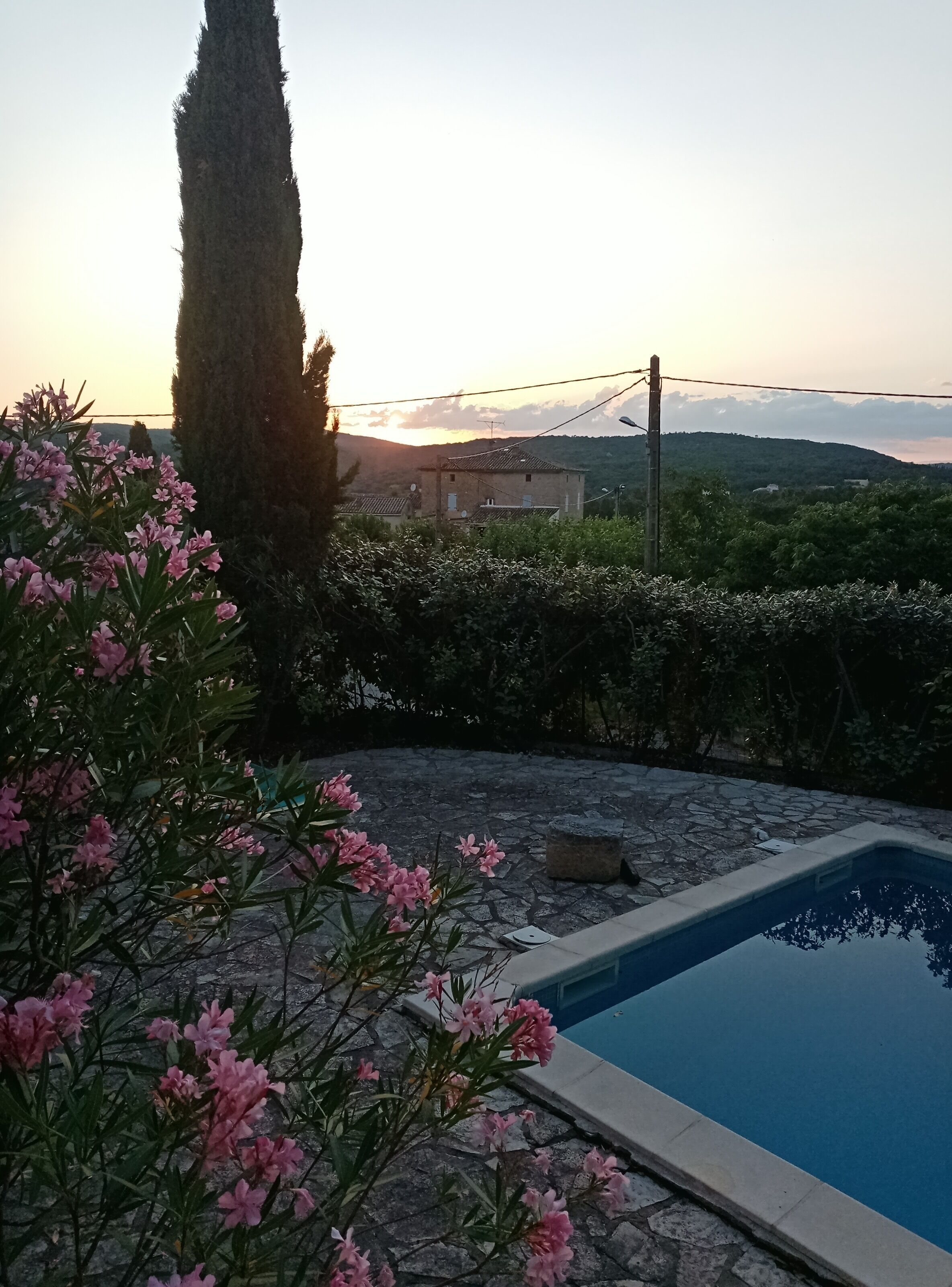 A swimming pool surrounded by stone tiles, with a view of a house and hills in the background.