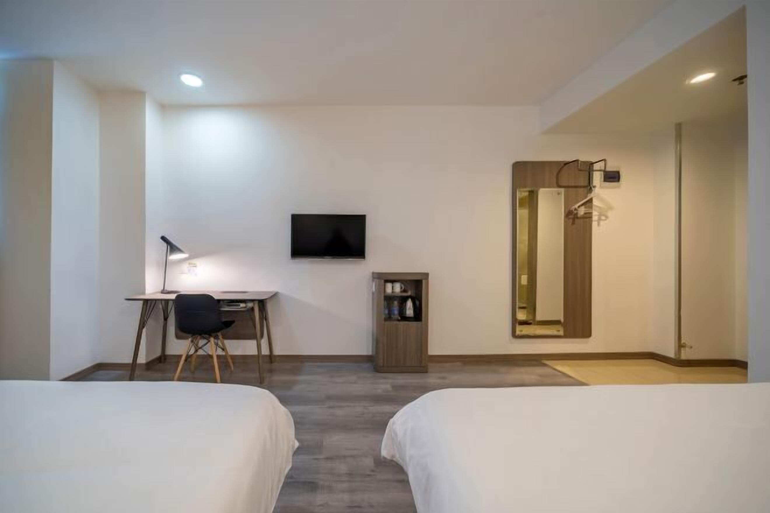 Quarto de hotel com duas camas, uma escrivaninha com cadeira, televisão na parede, um armário pequeno e um espelho com suporte para toalhas.