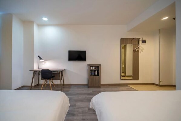 A hotel room with two beds, a desk with a chair, a TV mounted on the wall, a small cabinet, and a mirror with a towel rack.