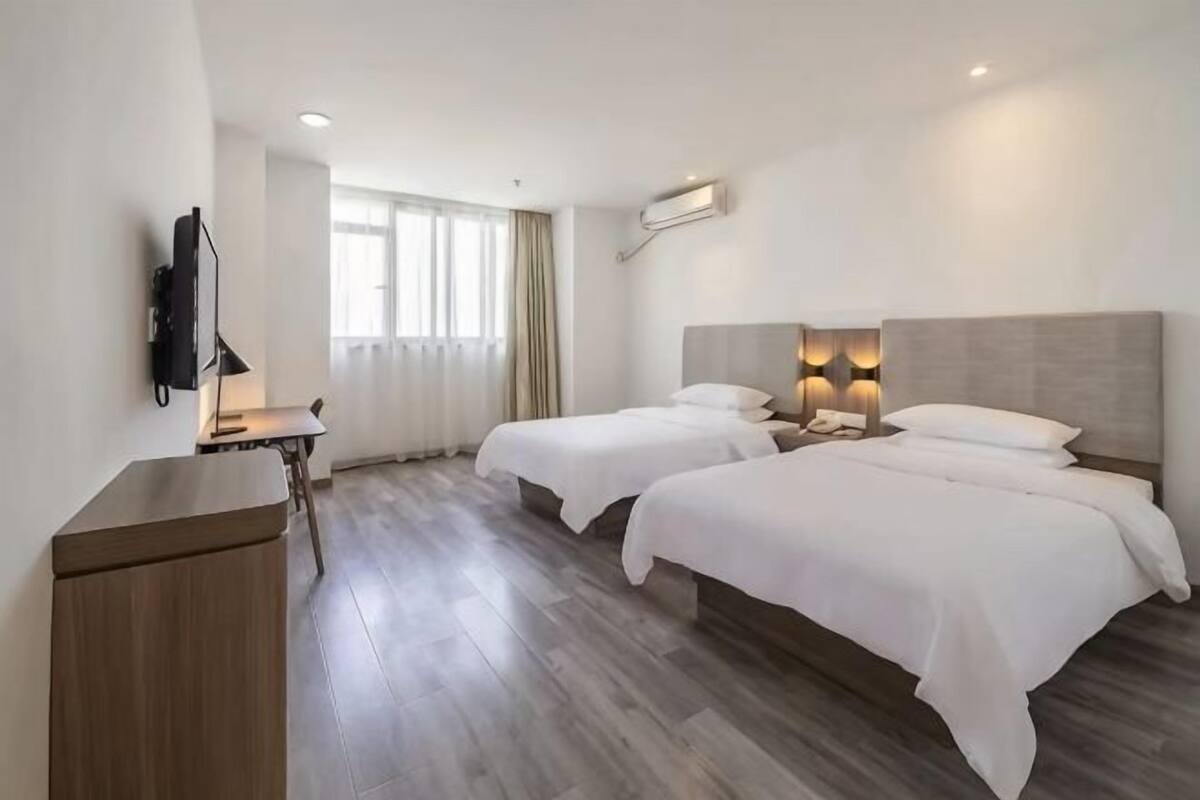 A hotel room with two beds, a desk with a lamp, a flat-screen TV, and a window with curtains.